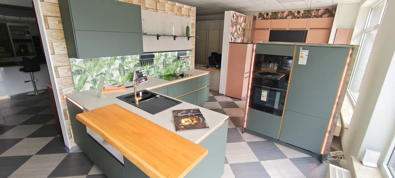 Mineralgrüne Nobilia Ausstellungsküche mit goldfarbenen Griffmulden, heller Arbeitsplatte und Geräteschrank